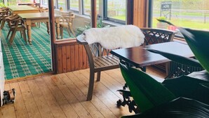 A room with wooden floors, a dining area with tables and chairs, and large windows offering a view of greenery and a sign for Invershin Hotel. - Invershin Hotel Bunkhouse & Bar (Invershin)
