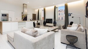 En moderne stue med sofa, sofabord og spiseområde med stole. Der er et køkkenområde med mikrobølgeovn og komfur. Værelset har store vinduer med gardiner og udsigt til bygninger.