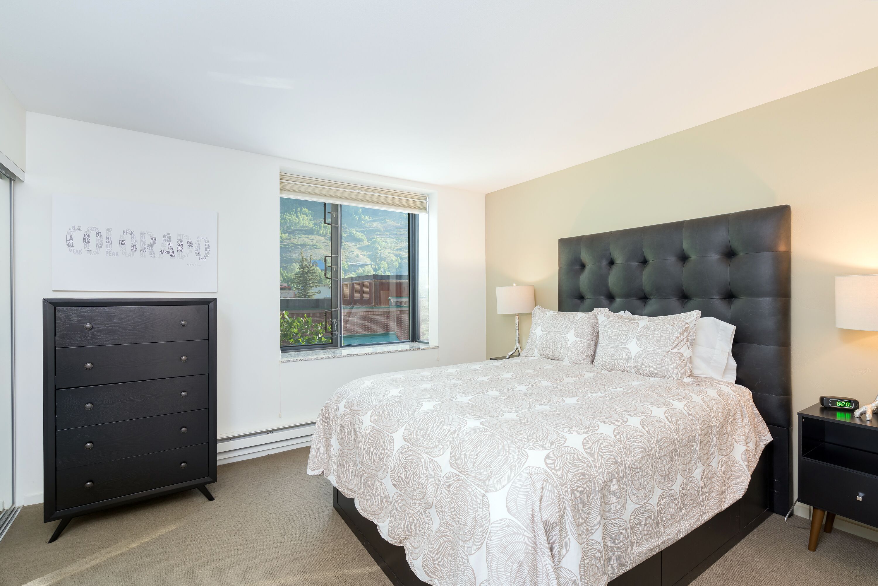 A bedroom with a large bed, a dresser, a nightstand, and a window with a view of greenery.
