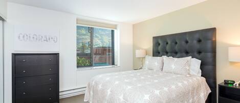 A bedroom with a large bed, a dresser, a nightstand, and a window with a view of greenery.