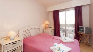 Habitación de hotel con cama, televisor, lámpara, mesita de mimbre y balcón con vistas.
