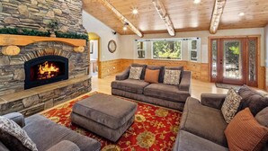A cozy living room with a stone fireplace, wooden ceiling beams, and a large window overlooking greenery.
