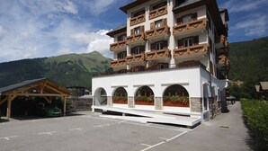 Ein mehrstöckiges Hotel mit Balkonen, einem überdachten Parkbereich und einer Bergkulisse.