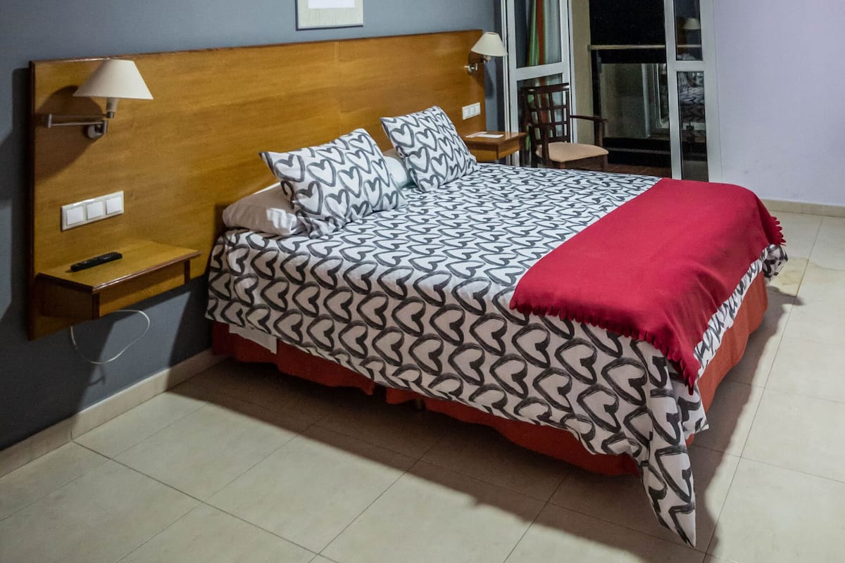 A hotel room with a bed, nightstand, lamp, and a view of the outside.