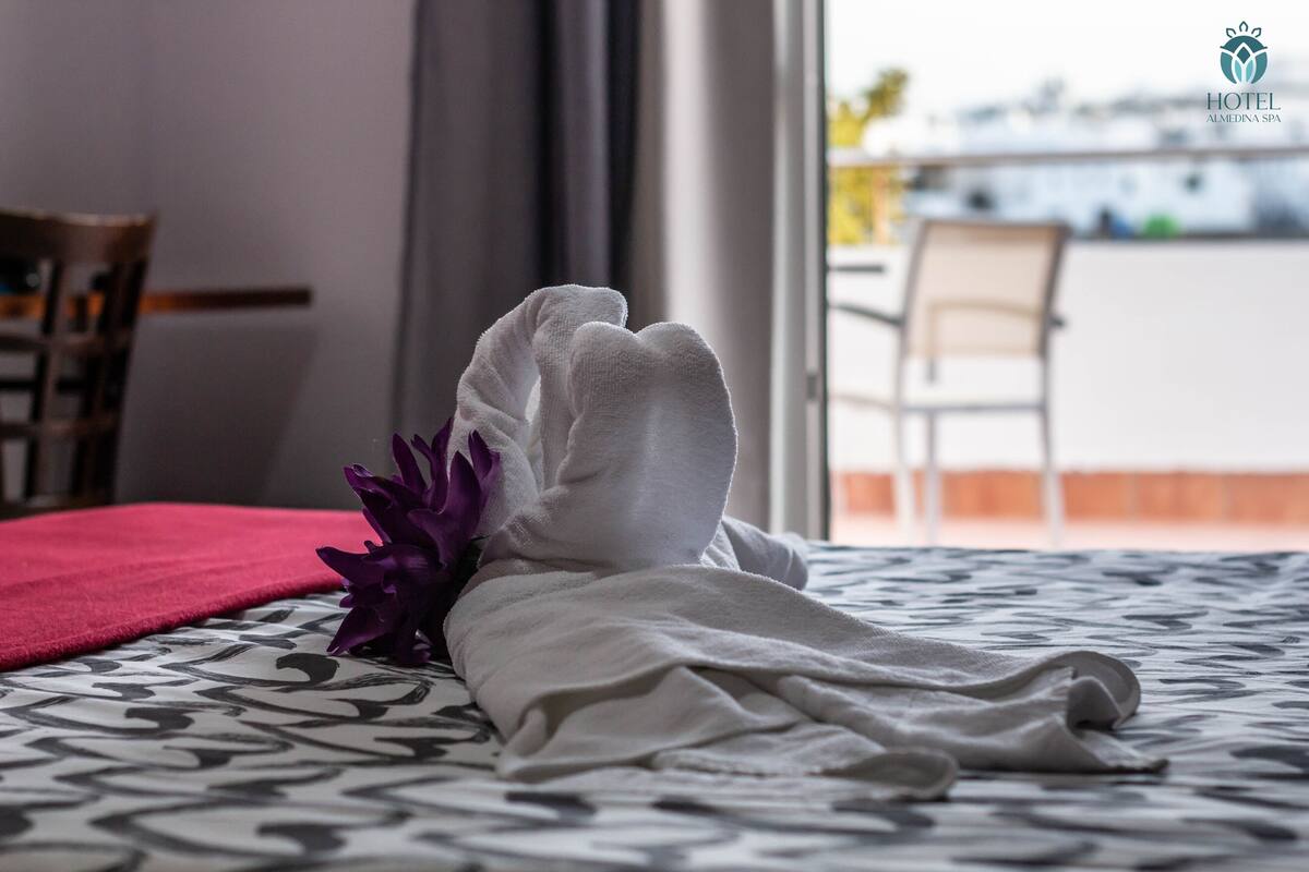 A neatly made bed with a white towel and purple flowers, a red blanket, and a patterned bedspread.