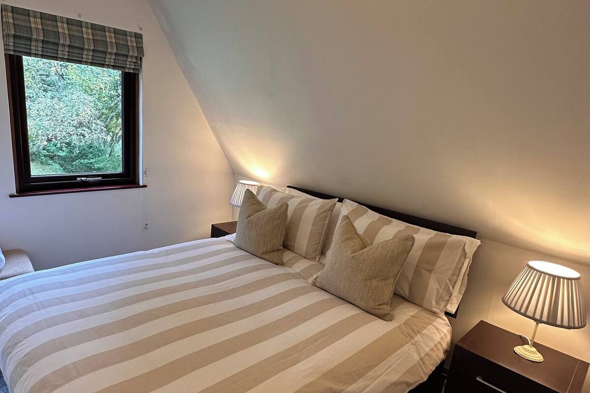 A bedroom with a bed, pillows, a nightstand, a lamp, and a window with a view of greenery.