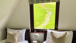 A room with two beds, a window with a view of a grassy area, a small bedside table with a lamp, and a rolled towel on the floor. - Glen Lodges (Rogart)
