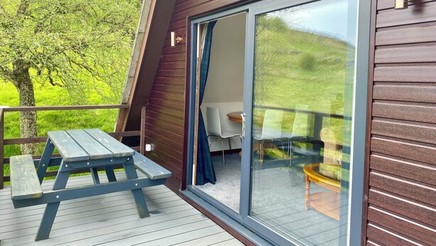 A wooden deck with a picnic table, a sliding glass door leading to an outdoor seating area with a table and chairs, and a view of a green landscape. - Glen Lodges (Rogart)