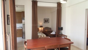 A compact hotel room with a wooden dining table, a single bed, a bedside table with a lamp, and a view of the kitchen area.
