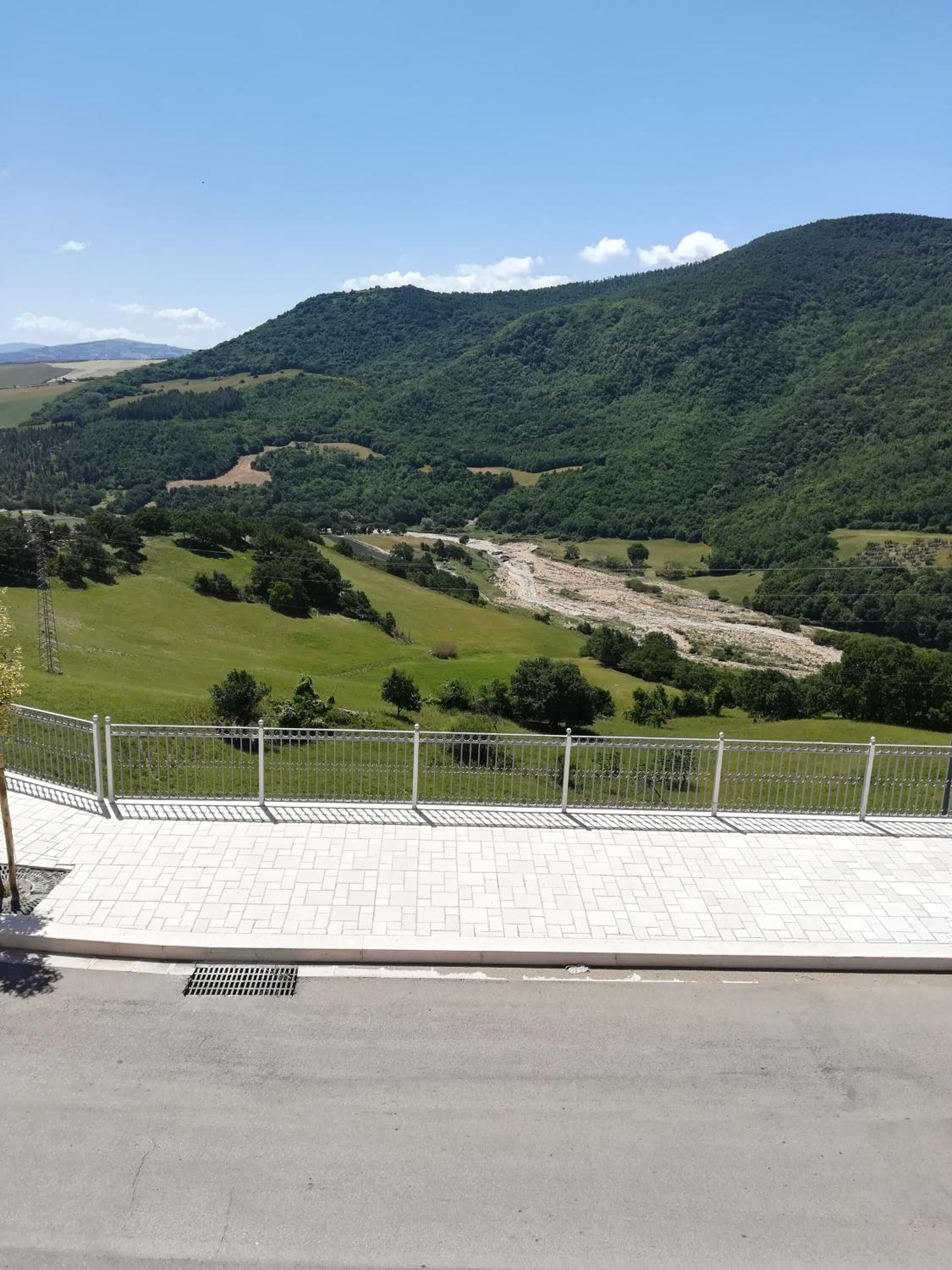 Eine gepflasterte Terrasse mit Metallgeländer, die auf ein Tal mit grünen Feldern und einem Fluss blickt.