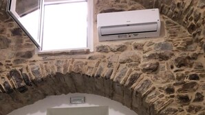 Una habitación con paredes de piedra, una puerta blanca, una ventana pequeña y un equipo de aire acondicionado sobre la puerta.