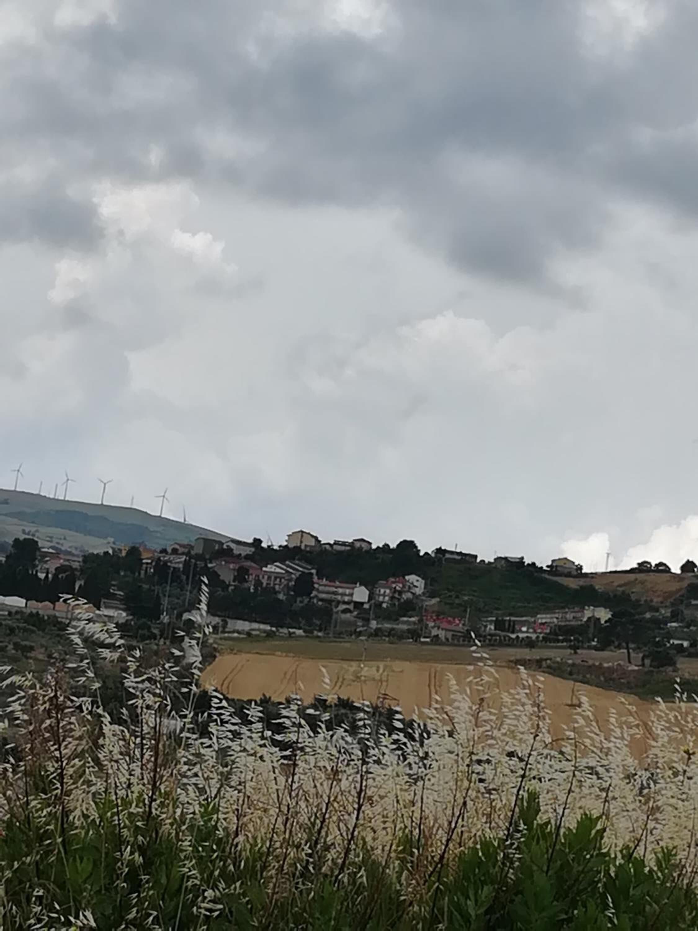 Un paisaje rural con aerogeneradores, un río y un pequeño asentamiento con casas y vegetación.