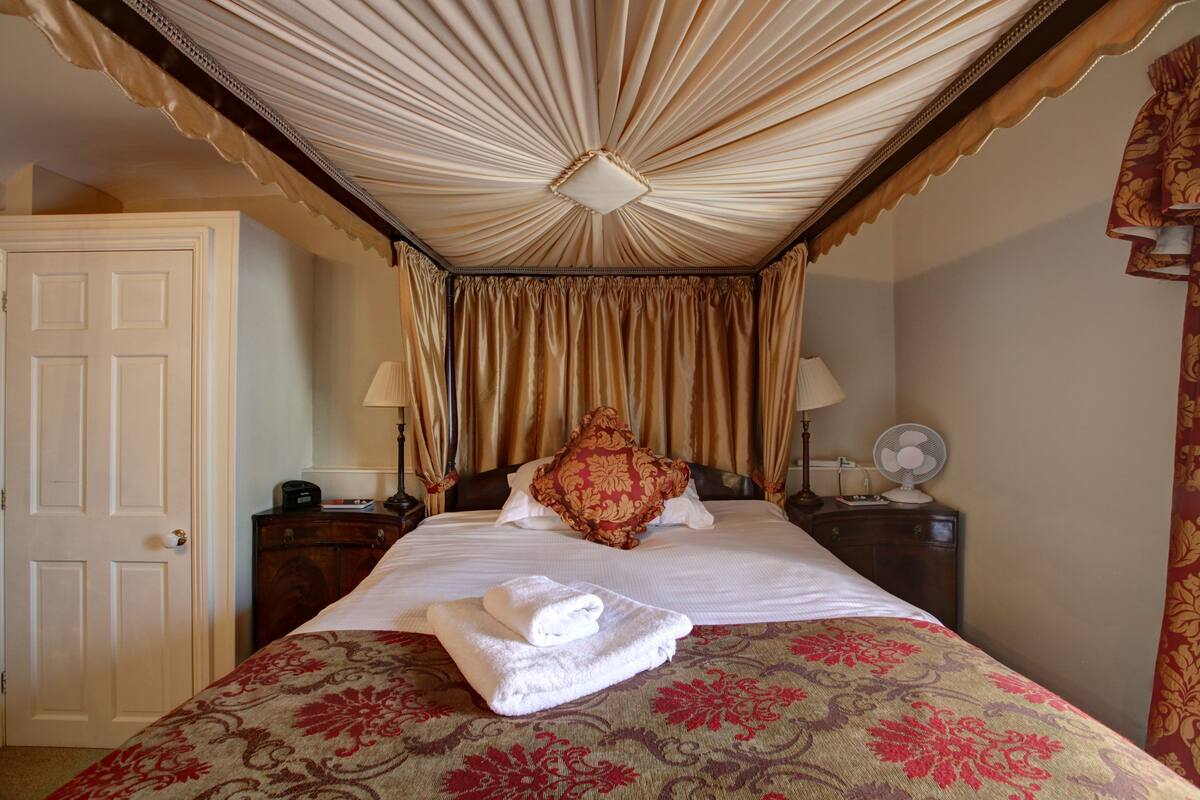 A hotel room with a bed, a bedside table with a lamp, a fan, and a door.