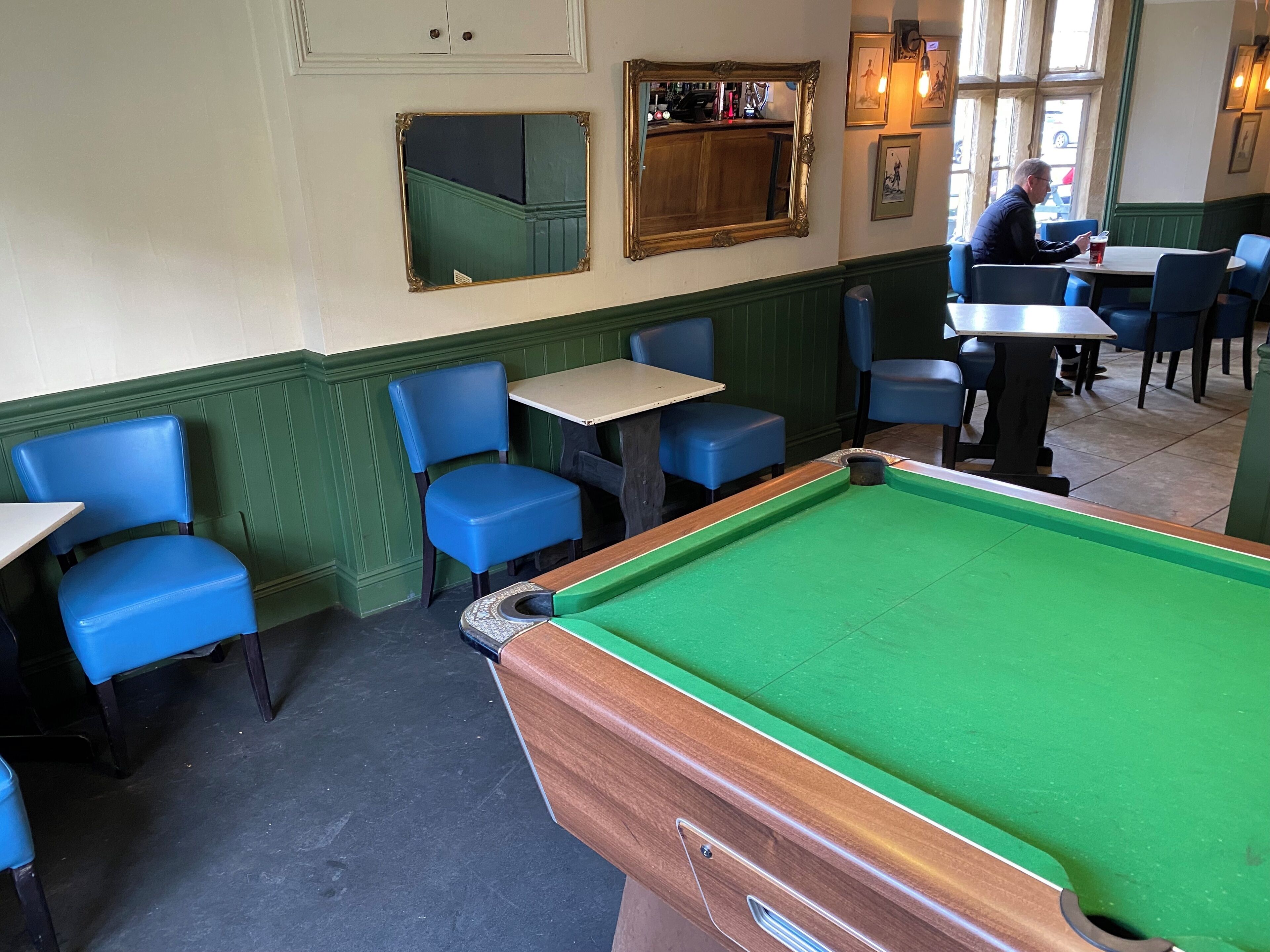 Een biljartruimte met groene tafels, blauwe stoelen en een bar met flessen.