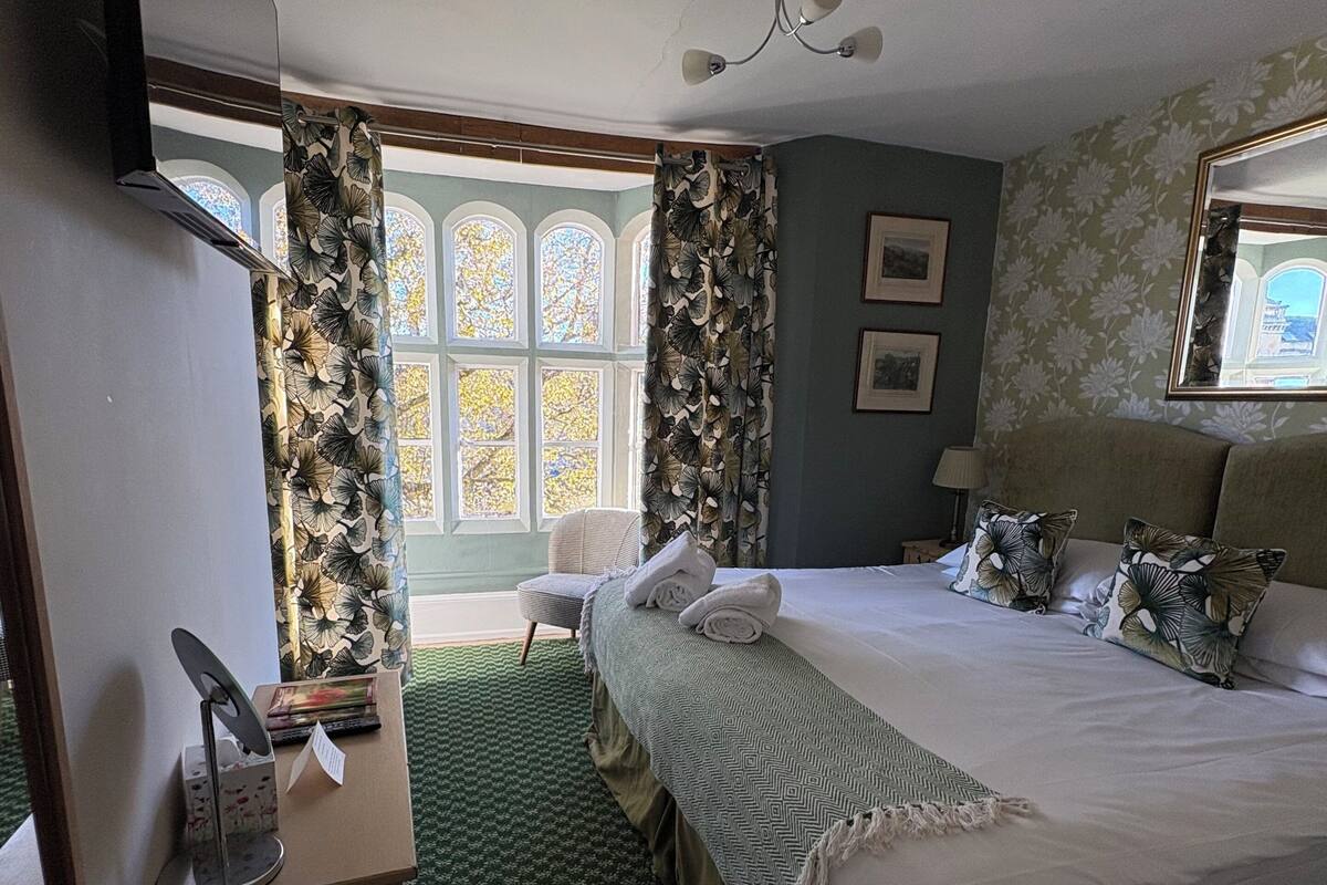 A bedroom with a bed, a desk, a chair, a window with curtains, and a wall-mounted lamp.