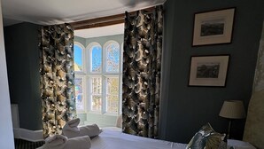 A hotel room with a bed, a window with curtains, a lamp, and two framed pictures on the wall. - The Swan Inn (Moreton-in-Marsh)