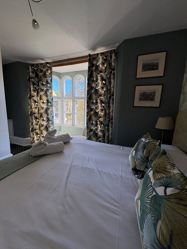 Habitación de hotel con cama, ventana con cortinas, lámpara y dos cuadros en la pared.