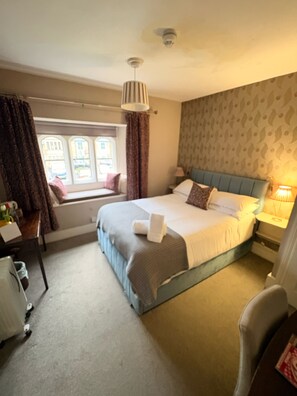 A bedroom with a bed, a window with curtains, a bedside table with a lamp, and a chair. - The Swan Inn (Moreton-in-Marsh)
