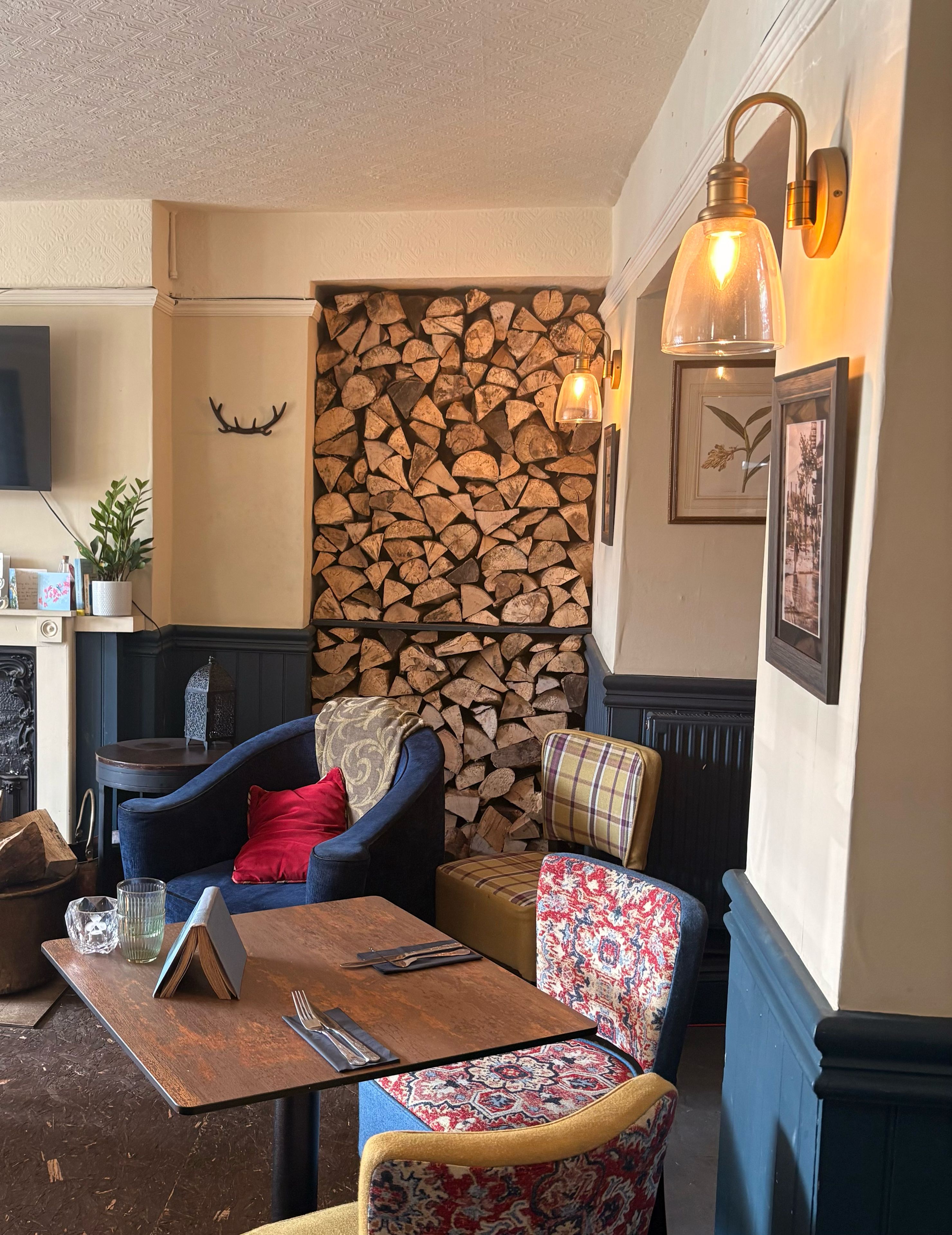 A cozy seating area with a fireplace, a wall of stacked logs, and a table with a book and glasses.