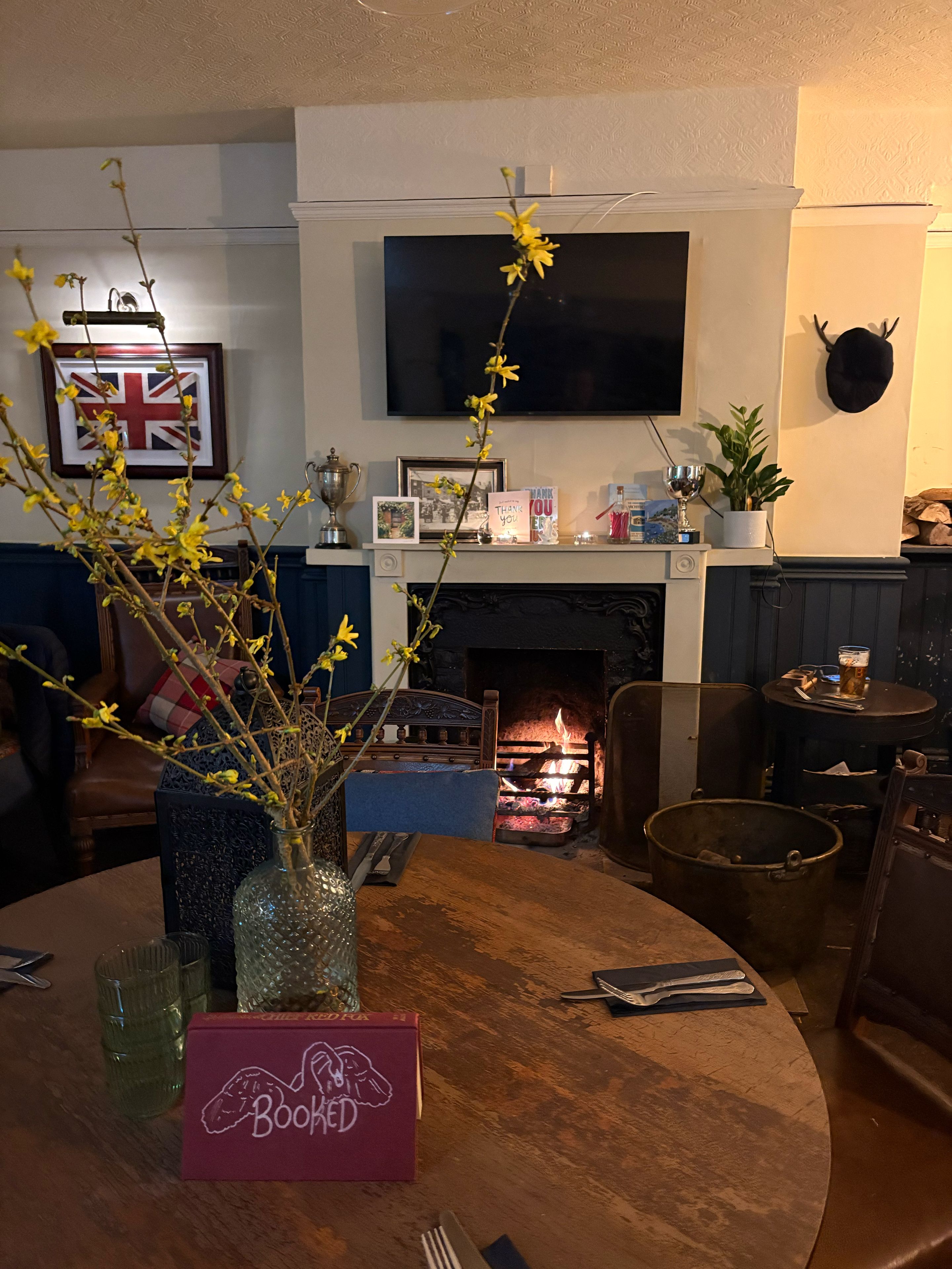 Un salon chaleureux avec une cheminée, un téléviseur fixé au mur et une table à manger agrémentée d’un vase de fleurs.