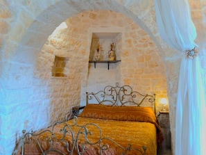 Une piĂšce avec un mur en pierre, un lit avec un cadre en mĂ©tal et une Ă©tagĂšre ornĂ©e dâobjets dĂ©coratifs.