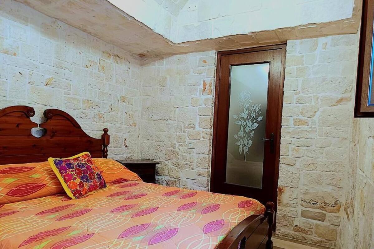 A bedroom with a stone wall, a wooden bed with a floral bedspread, a wooden door with a frosted glass panel, and a small nightstand.