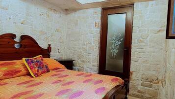 A bedroom with a stone wall, a wooden bed with a floral bedspread, a wooden door with a frosted glass panel, and a small nightstand.