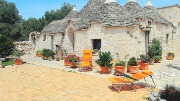 A stone building with multiple conical roofs, a paved courtyard with orange chairs, and potted plants.