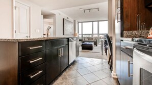 A modern kitchen with dark cabinetry, a granite countertop, and a view to a living area with a large window. - Bluebird Suites in Pentagon City (Arlington)