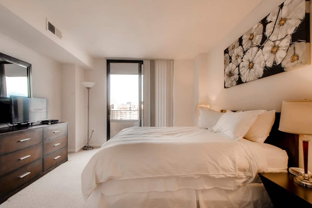 A hotel room with a bed, a dresser, a television, a painting, and a view of the city.