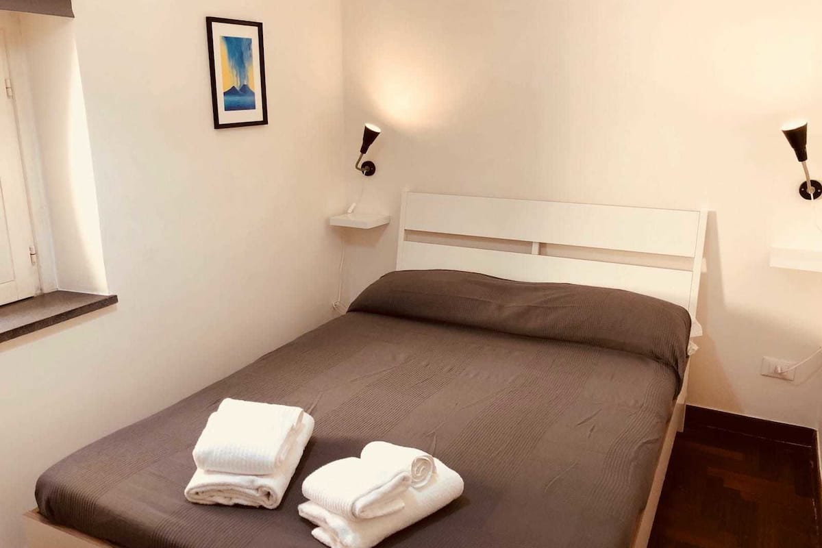 A neatly made bed with two folded white towels, a framed picture on the wall, and two wall-mounted lamps.