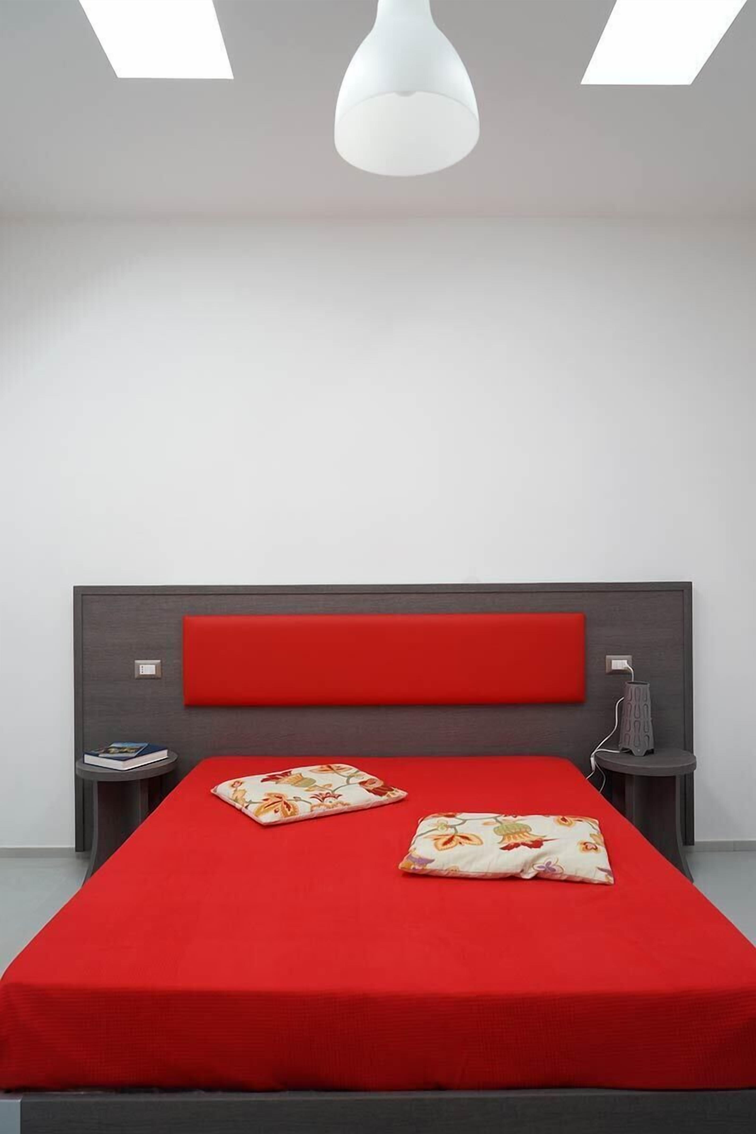 Een moderne slaapkamer met een rode spreideken, twee kussens, een nachtkastje met een telefoon en een wandlamp.