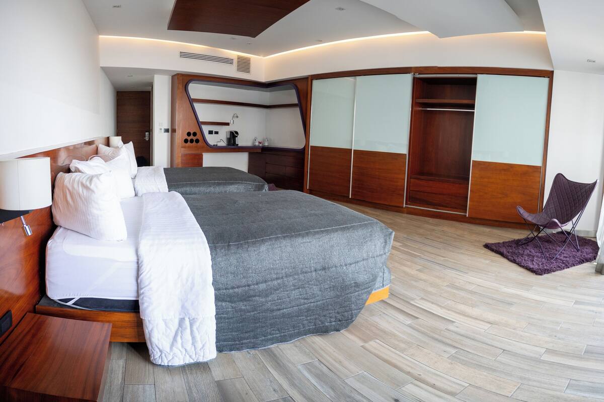 A modern hotel room with two beds, a wooden floor, a seating area with a purple rug, and a wardrobe with sliding doors.