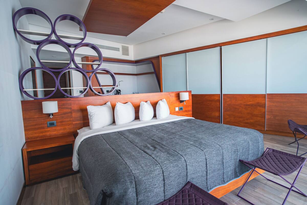 A modern hotel room with a large bed, a wooden headboard, and a seating area with a chair.