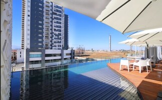 Un hotel moderno con piscina infinita en la azotea, vista a un edificio alto y mobiliario exterior.