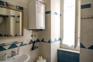 A bathroom with a white sink, a glass shelf, a mirror, a towel rack, a shower area, and a window with curtains. - GuestHouse Marcanto - Duomo (Naples)