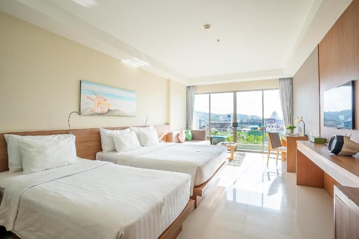 A hotel room with two beds, a desk, a chair, a TV, and a large window with a city view.