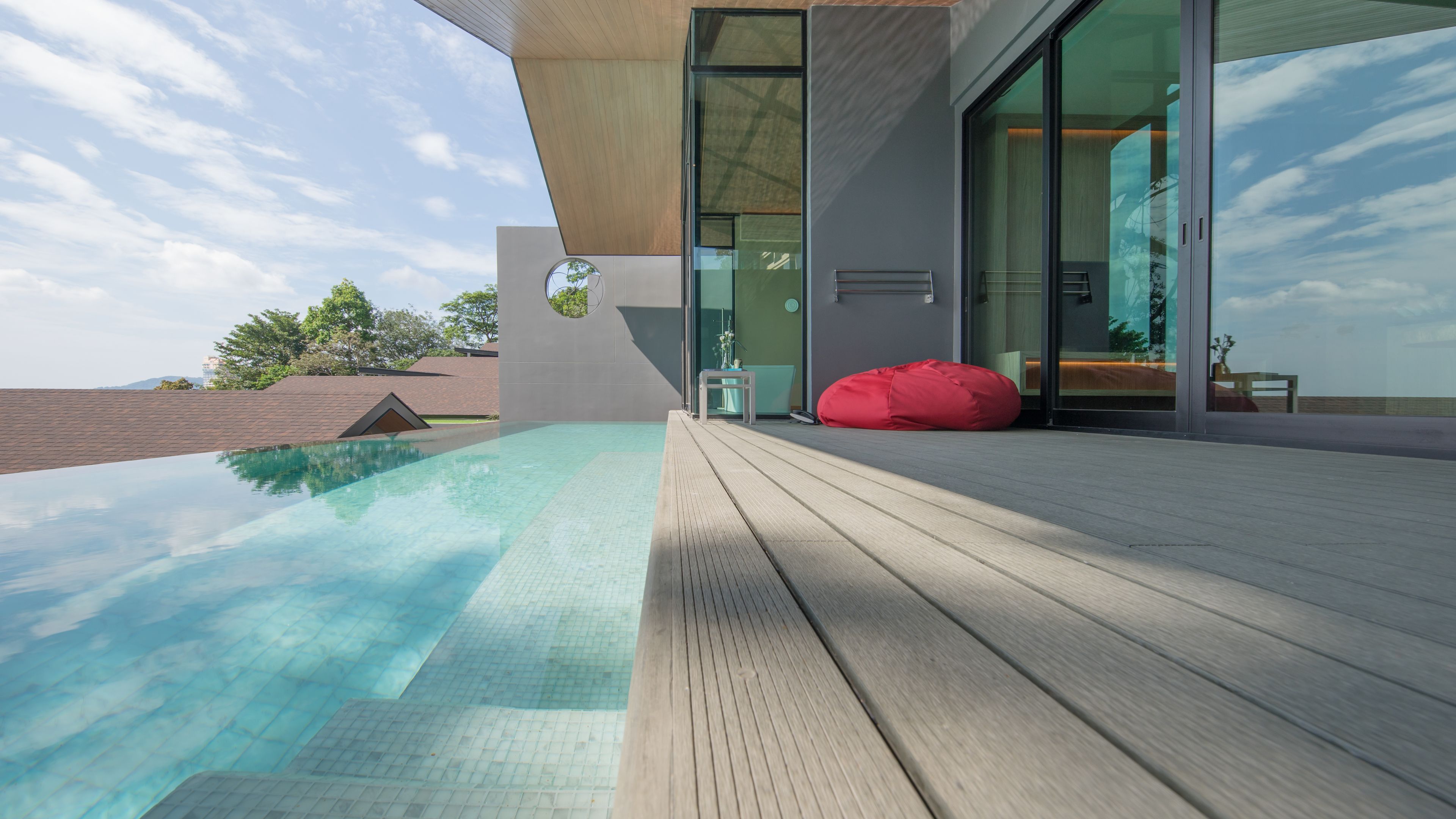 Un área de piscina moderna al aire libre con una terraza de madera, un salón con paredes de cristal y un sillón de pie rojo.