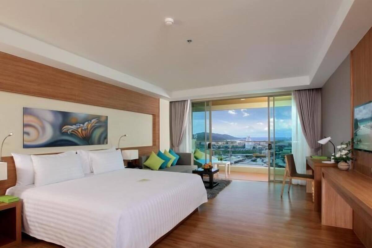 A hotel room with a large bed, a desk, a chair, a TV, and a city view through large windows.