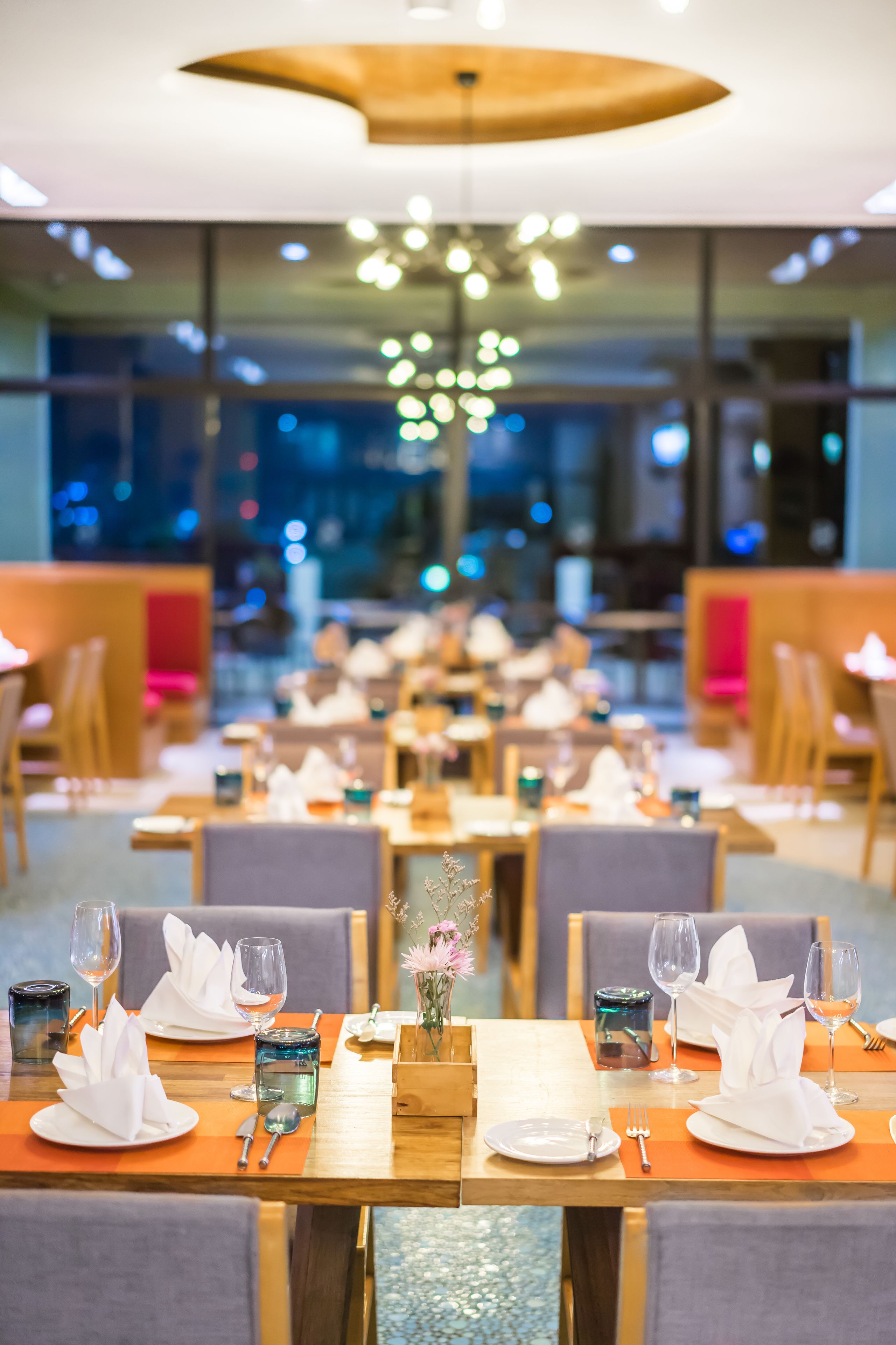 Um restaurante com mesas preparadas para refeições, com um lustre moderno e vista para a cidade à noite.