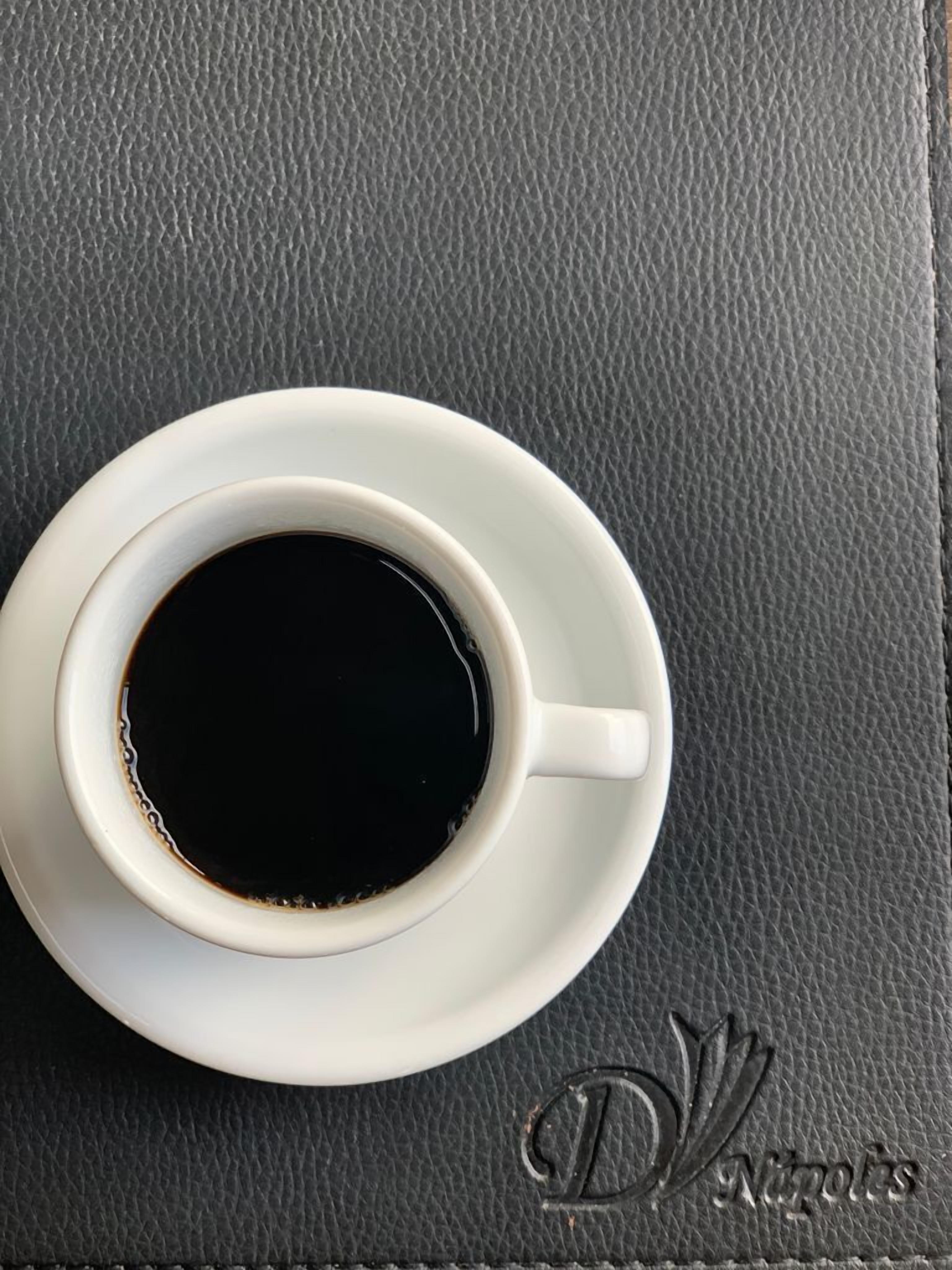 Una taza blanca de café negro sobre un platito blanco, colocada sobre una superficie negra texturizada.