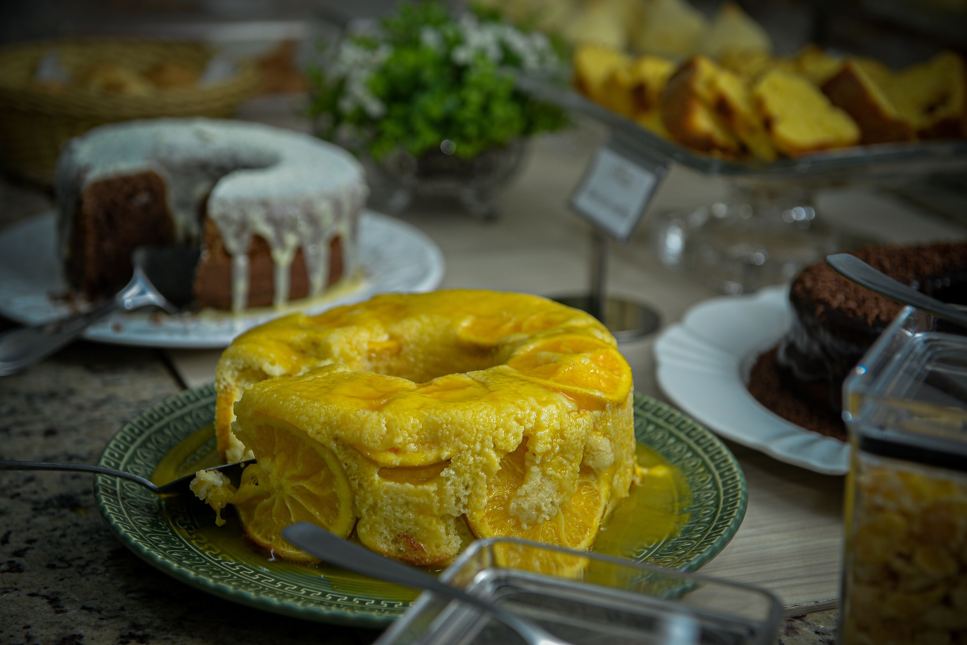 Variedad de pasteles expuestos, incluyendo un pastel amarillo con rodajas de naranja encima.