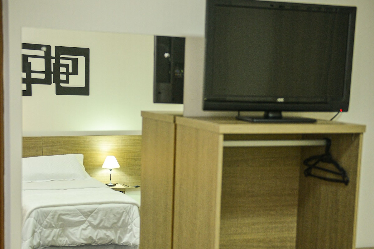 Ein Hotelzimmer mit Bett, Fernseher auf einem Holzständer, Nachttisch mit Lampe und Kleiderschrank mit Kleiderhaken.