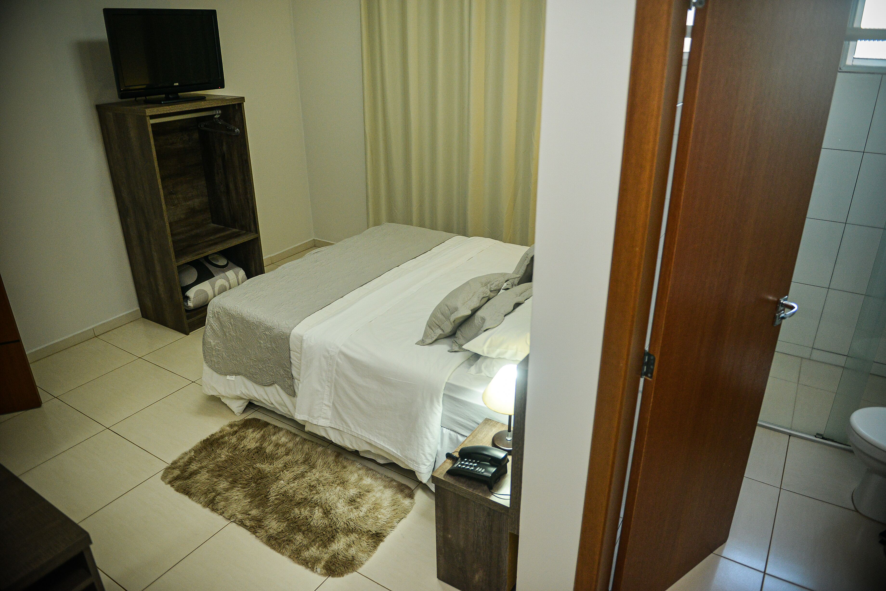 A bedroom with a bed, a wooden wardrobe, a television on a stand, a bathroom with a toilet, and a door leading to another room.