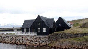 A black wooden house with a white porch, situated by a rocky shoreline with a body of water in the background. - Sunnuberg Guesthouse (Skagafjörour)