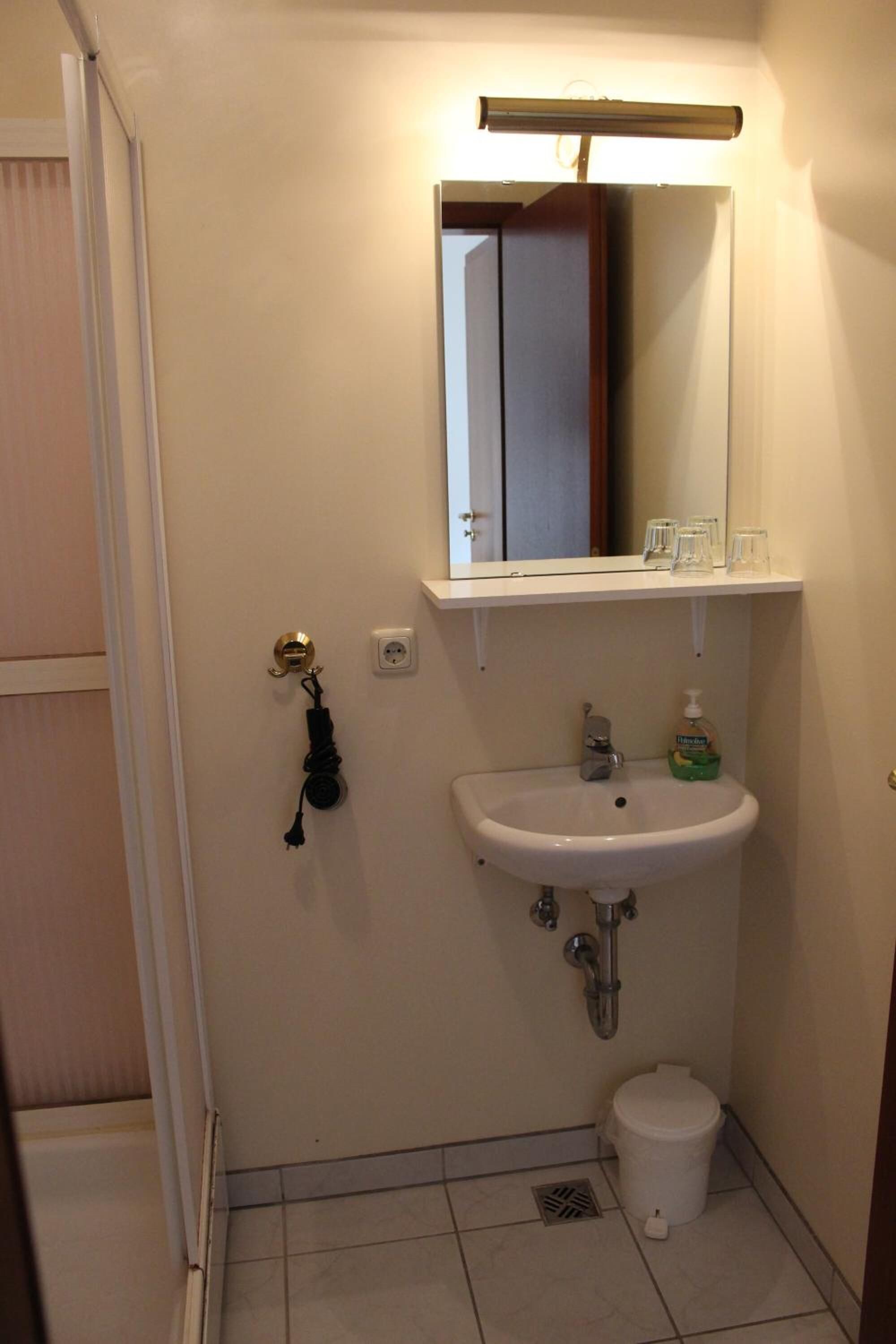 Een compacte badkamer met een witte wastafel, een spiegel en een toilet. Boven de wastafel bevindt zich een klein schapje met enkele glazen.