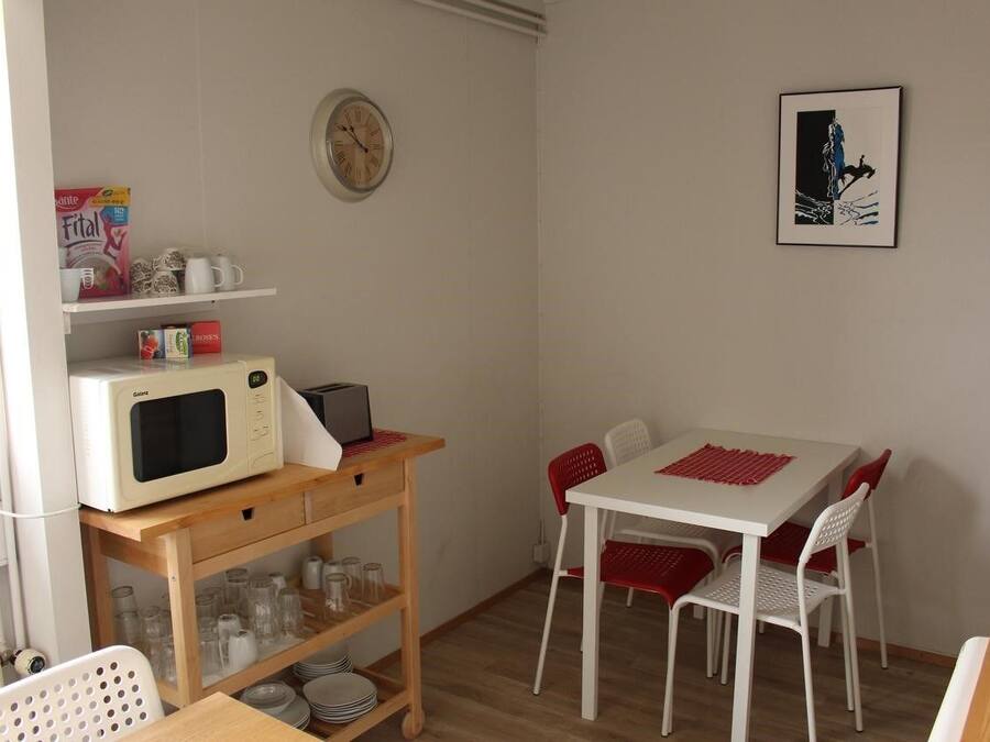 Een compacte keuken met een magnetron, broodrooster en koffiezetapparaat op een houten plank. Een eettafel met rode stoelen en een lijst met een afbeelding aan de muur.