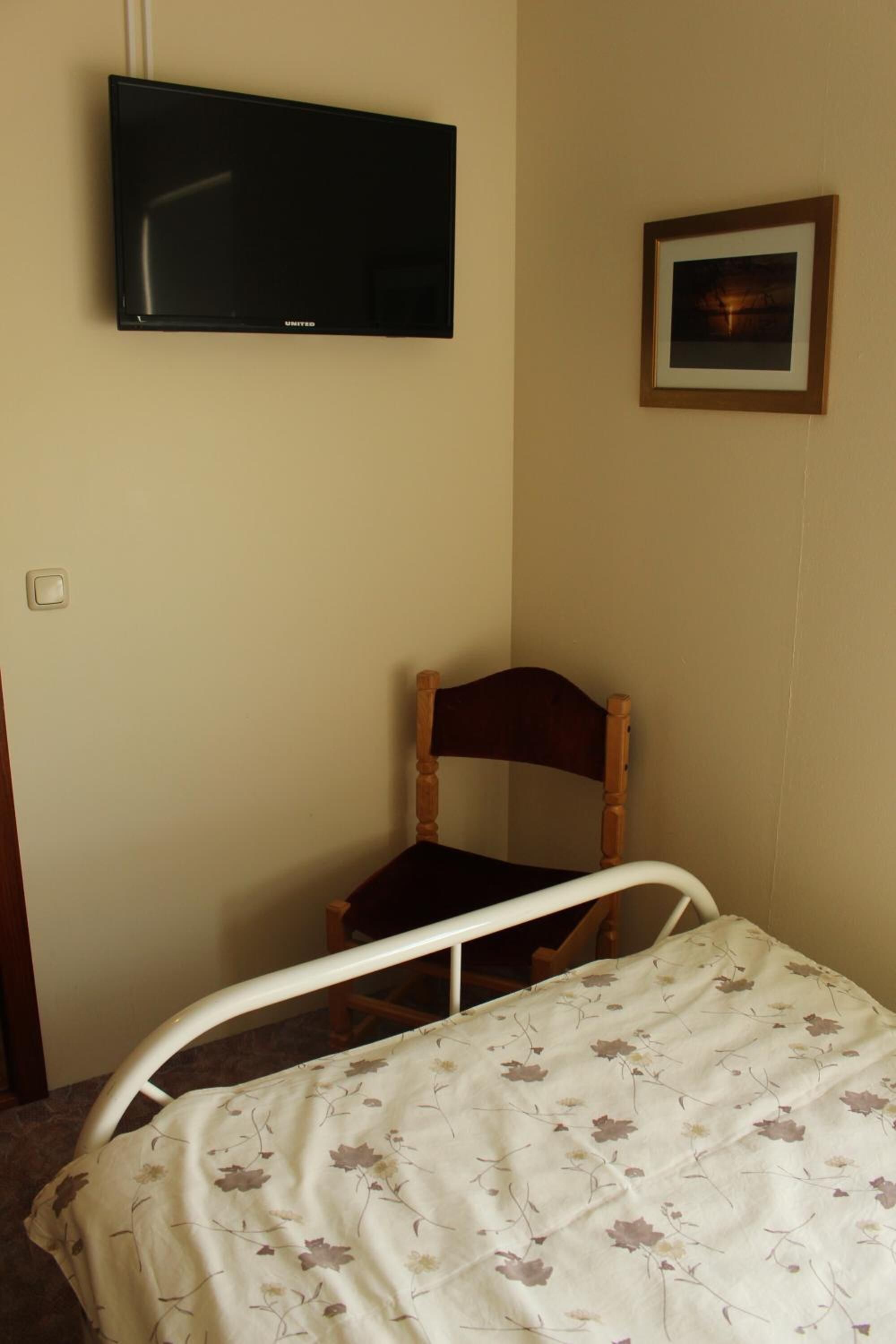Une chambre à coucher avec un lit, une chaise en bois, un téléviseur fixé au mur et un tableau encadré.