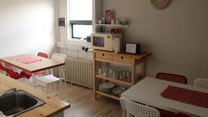 A compact kitchen with a microwave, a small refrigerator, and a window with blinds. There are white chairs and a table with a red placemat. - Sunnuberg Guesthouse (Skagafjörour)