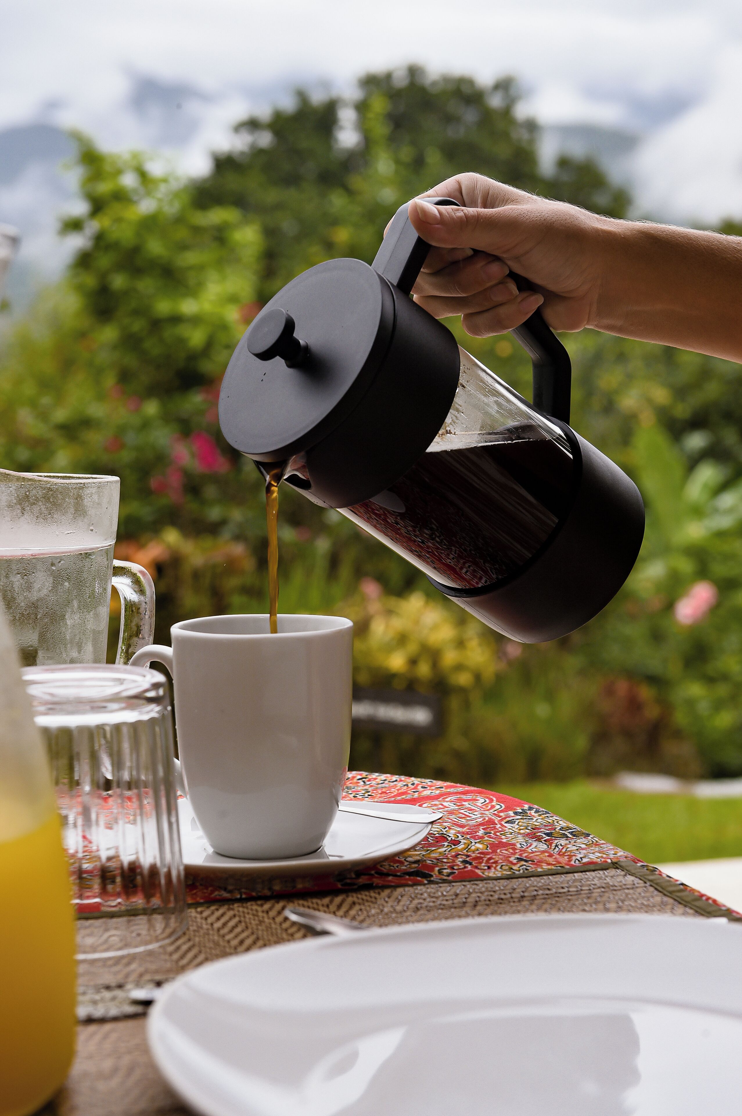 A hand pouring coffee into a white cup, with a glass of water and a glass of orange juice on a table.